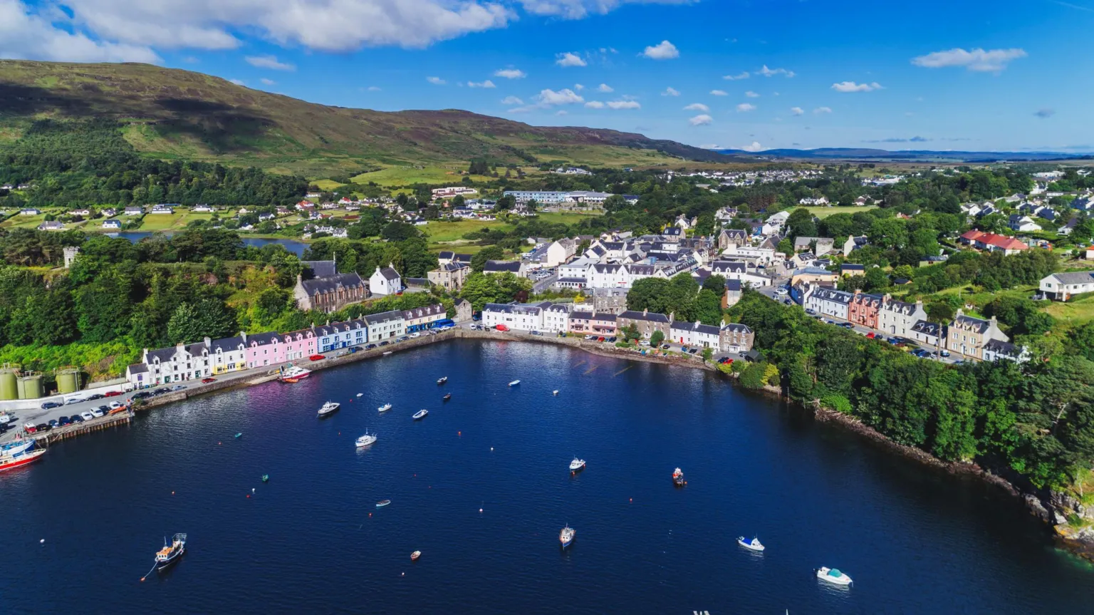  An aerial view over a bay filled with small boats to Portree on Skye. The town has houses and other buildings among trees. Some of the houses have colourful exterior walls, including pink and blue. A green hill rises behind the house. It is a sunny day with white clouds in a blue sky.