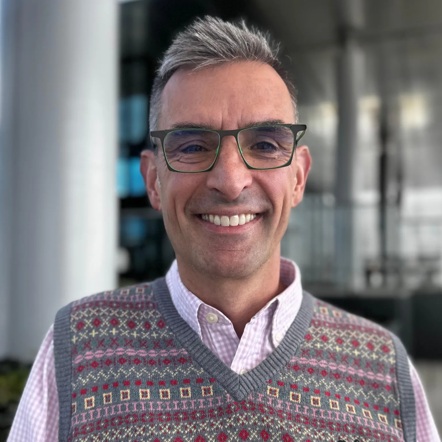 Simon Blake, who is wearing a pink short, a sleeveless knitted jumper and glasses. He has short grey hair and is smiling.