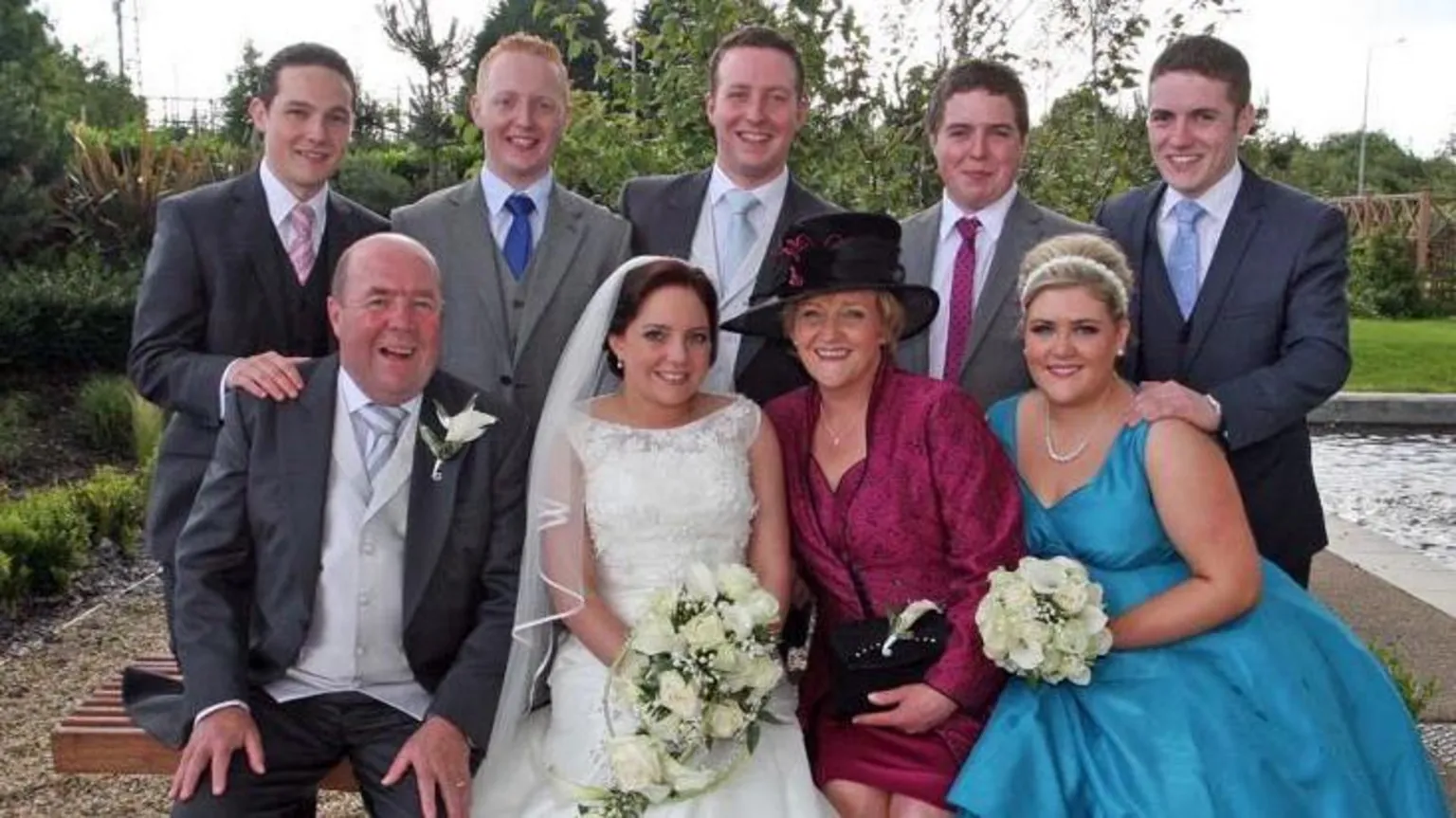 BELL FAMILY The Bell family of nine - two parents and their seven adult children - pose for a wedding photo. The parents are sitting on either side of their daughter, the bride who is wearing a white lace dress and a veil. The men are all in formal suits.