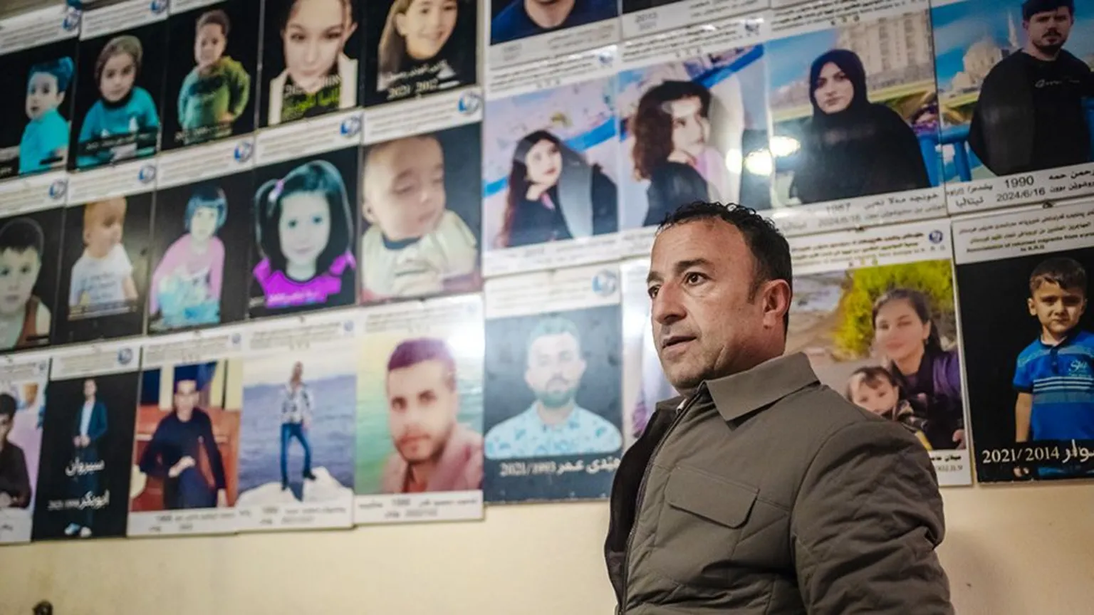 An indoor space with a wall densely covered in rows of printed portrait photographs, each photo mounted on a card with lines of text beneath it, some written in Arabic numerals and script. The photographs include adults and children shown against varied backgrounds, arranged in a grid that fills most of the wall from left to right. In the foreground, Bakr Ali stands slightly turned to the side, wearing a padded, zip-up jacket. He is positioned to the right of the frame and appears to be looking toward the wall of photographs.
