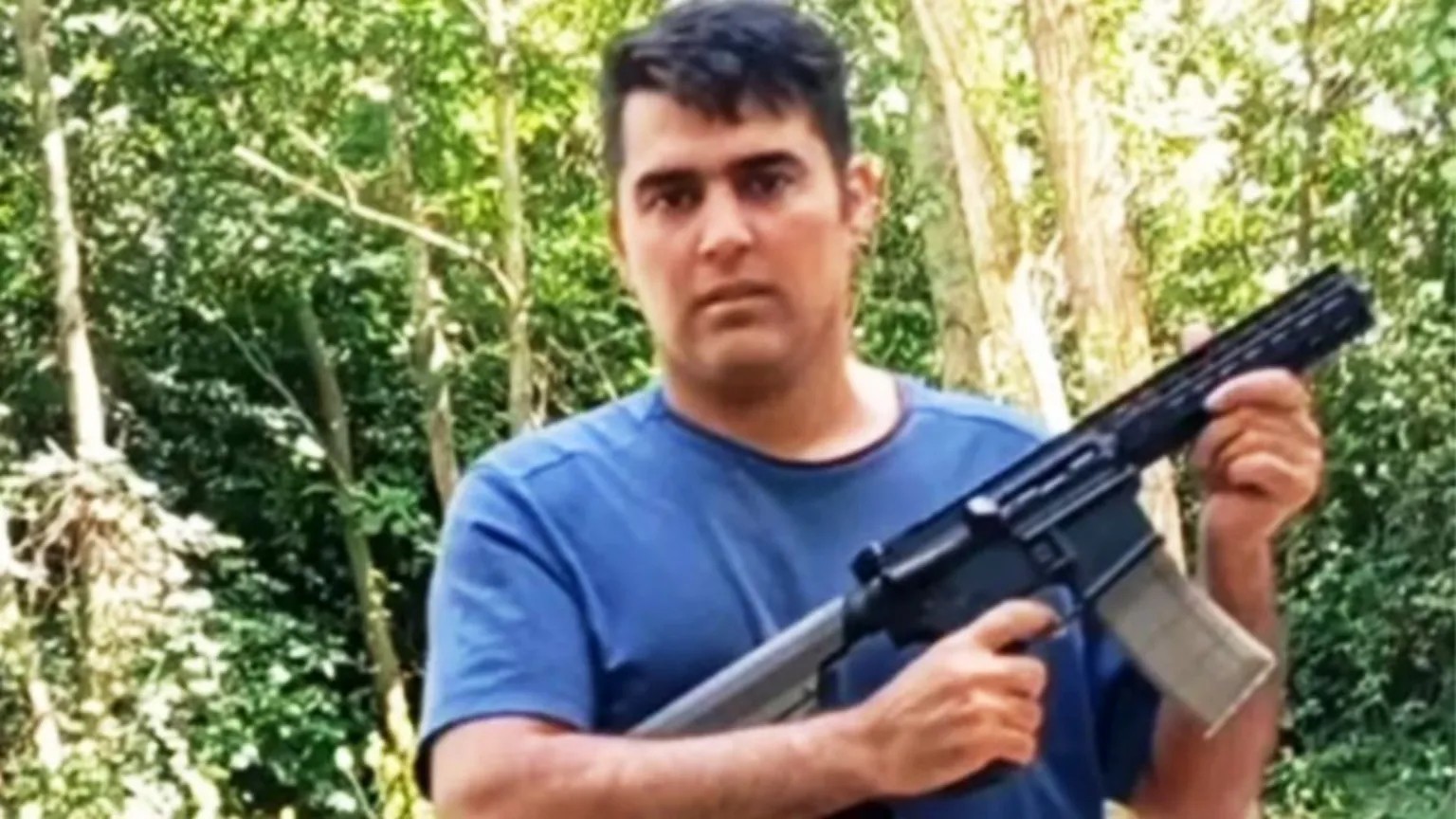 Noah Aaron stands outdoors in a wooded area with dense green trees and sunlight filtering through the foliage. He is wearing a short-sleeved blue T-shirt and is holding a long firearm with both hands; the firearm has a visible magazine and is angled slightly across the body. The background consists of tree trunks, leaves, and undergrowth, with no buildings or other people visible.