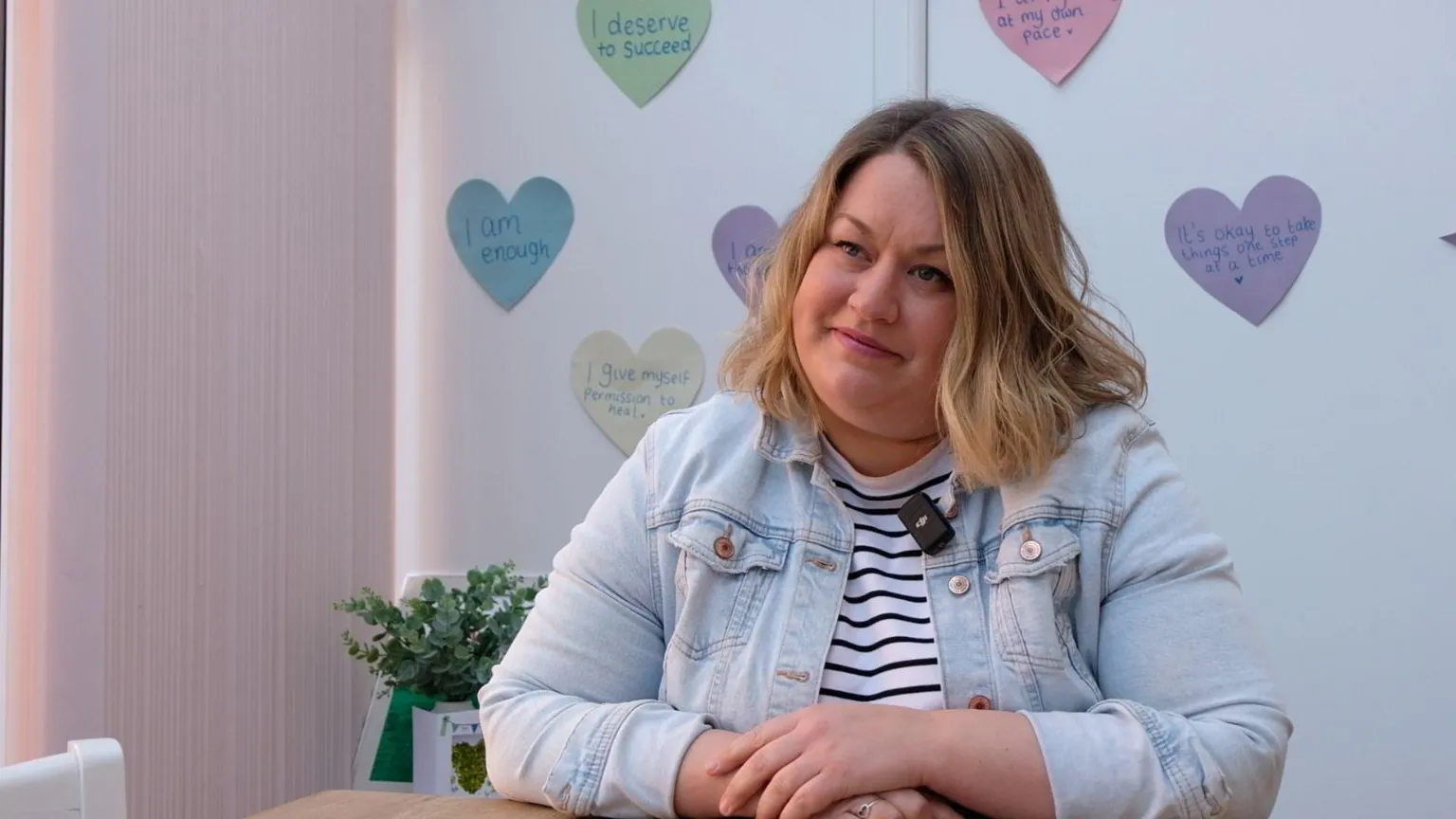 Alabaré Helen Brian has dark blonde shoulder length hair and is sat wearing a light blue denim jacket with her arms folded in front of her on a table. Behind her there is a plant and also some post-it notes on a wall displaying positive affirmations such as 