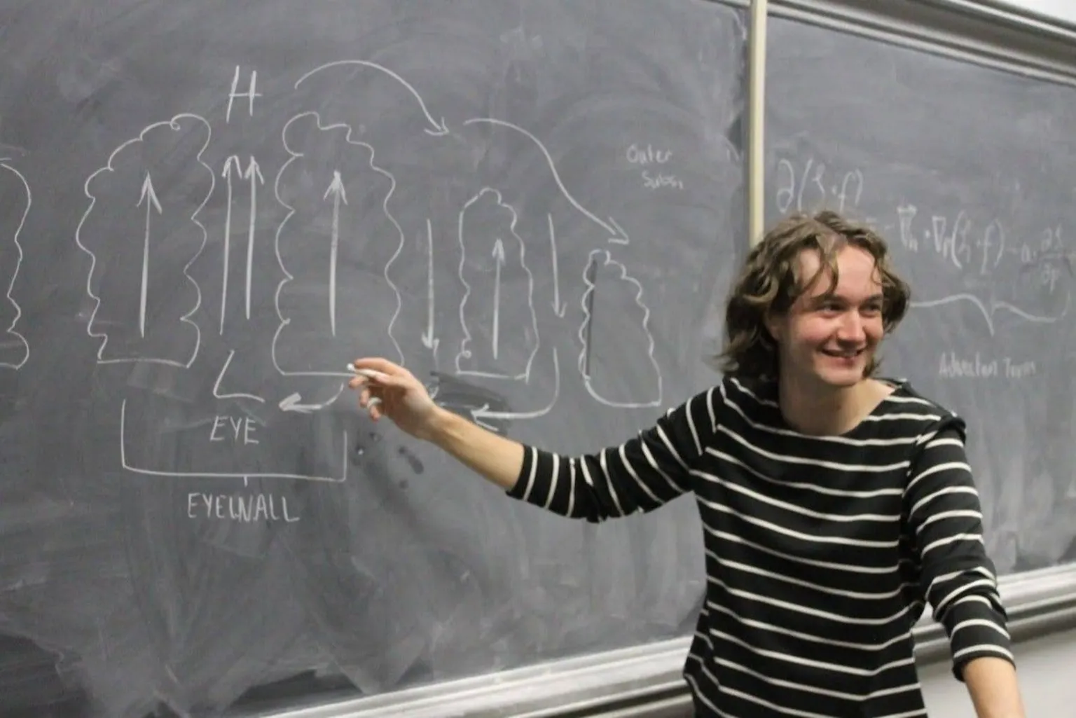 Courtesy of Aubrey Palmer Meteorology student Aubrey Palmer points to a weather diagram on a blackboard with a piece of chalk while smiling and facing a classroom. 
