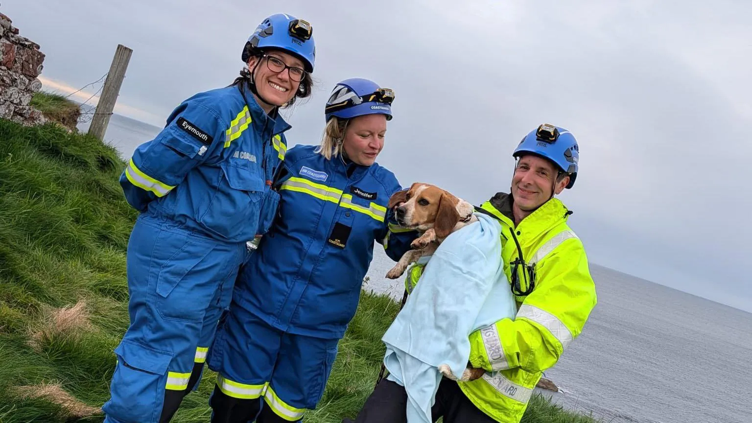 Eyemouth Coastguard Rescue Team Two people in blue outdoor wear beside a man in a high vis jacket who is holding a dog wrapped in a blanket
