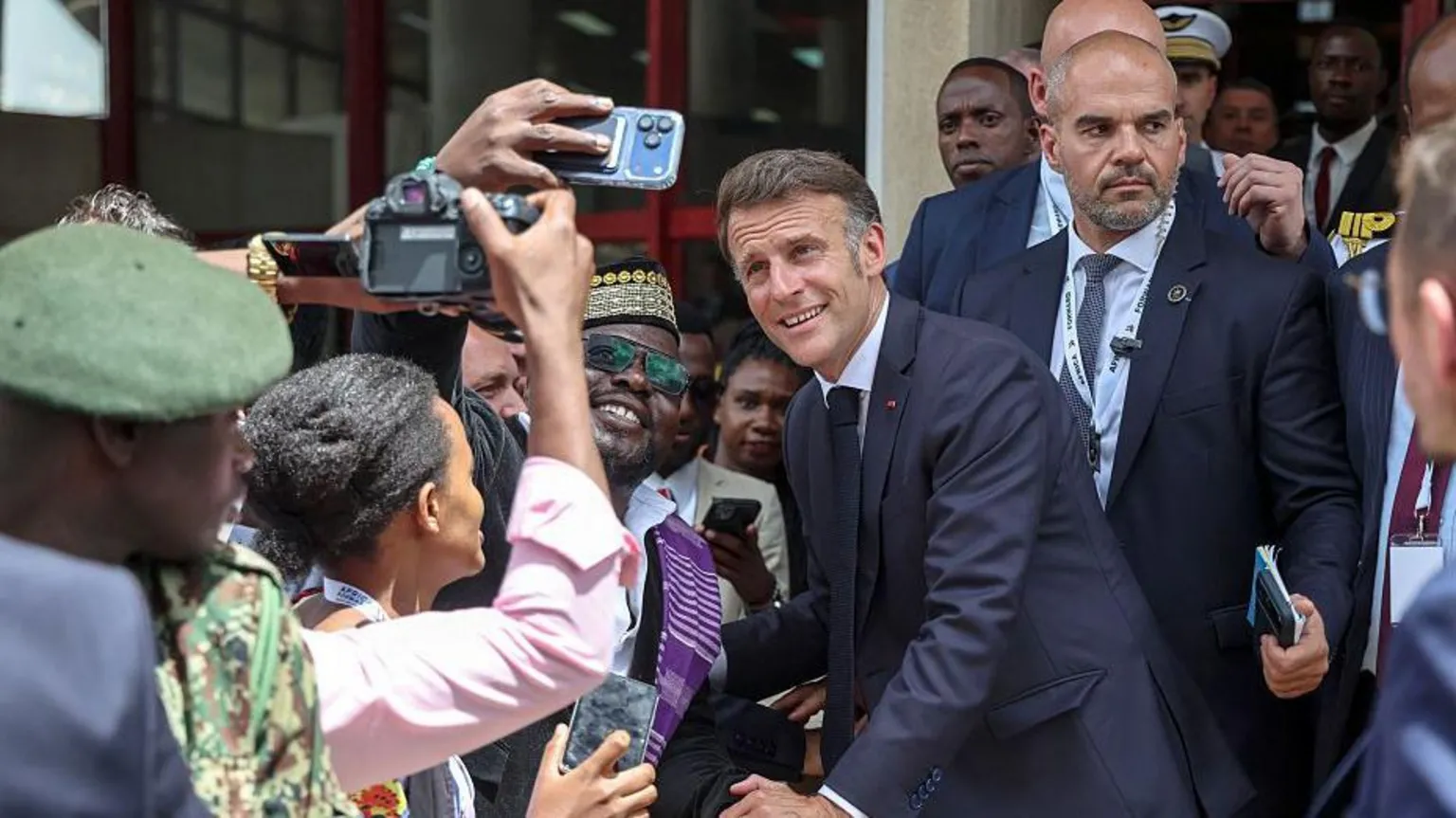 AFP via Macron posing for a selfie as a man smiles and holds up his mobile phone. Others near them are holsing cameras and behind Macron is his security. Macron and his entourage are walking out of a building.