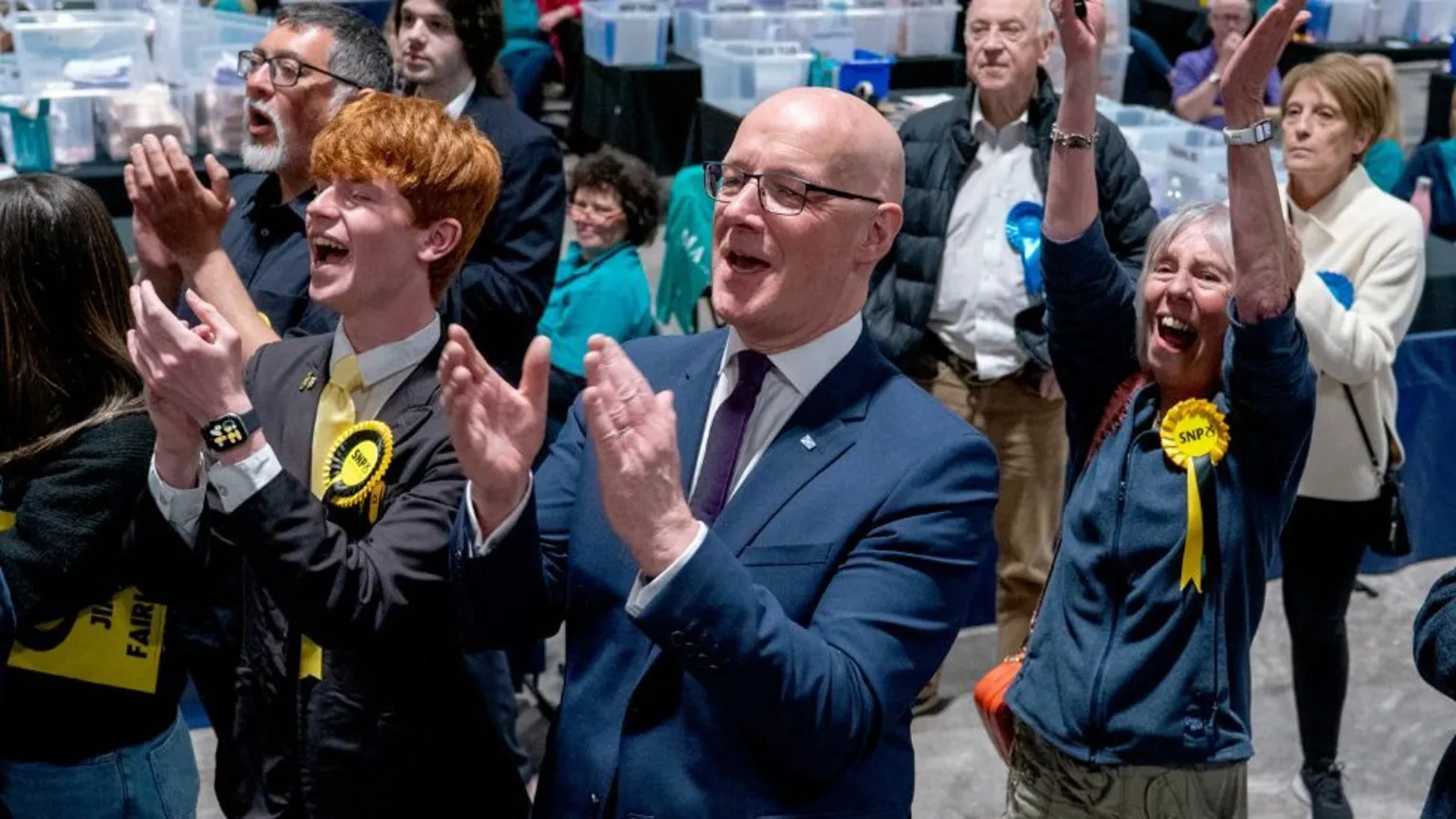  John Swinney, who is bald with glasses, claps with celebrating SNP supporters at an election count 