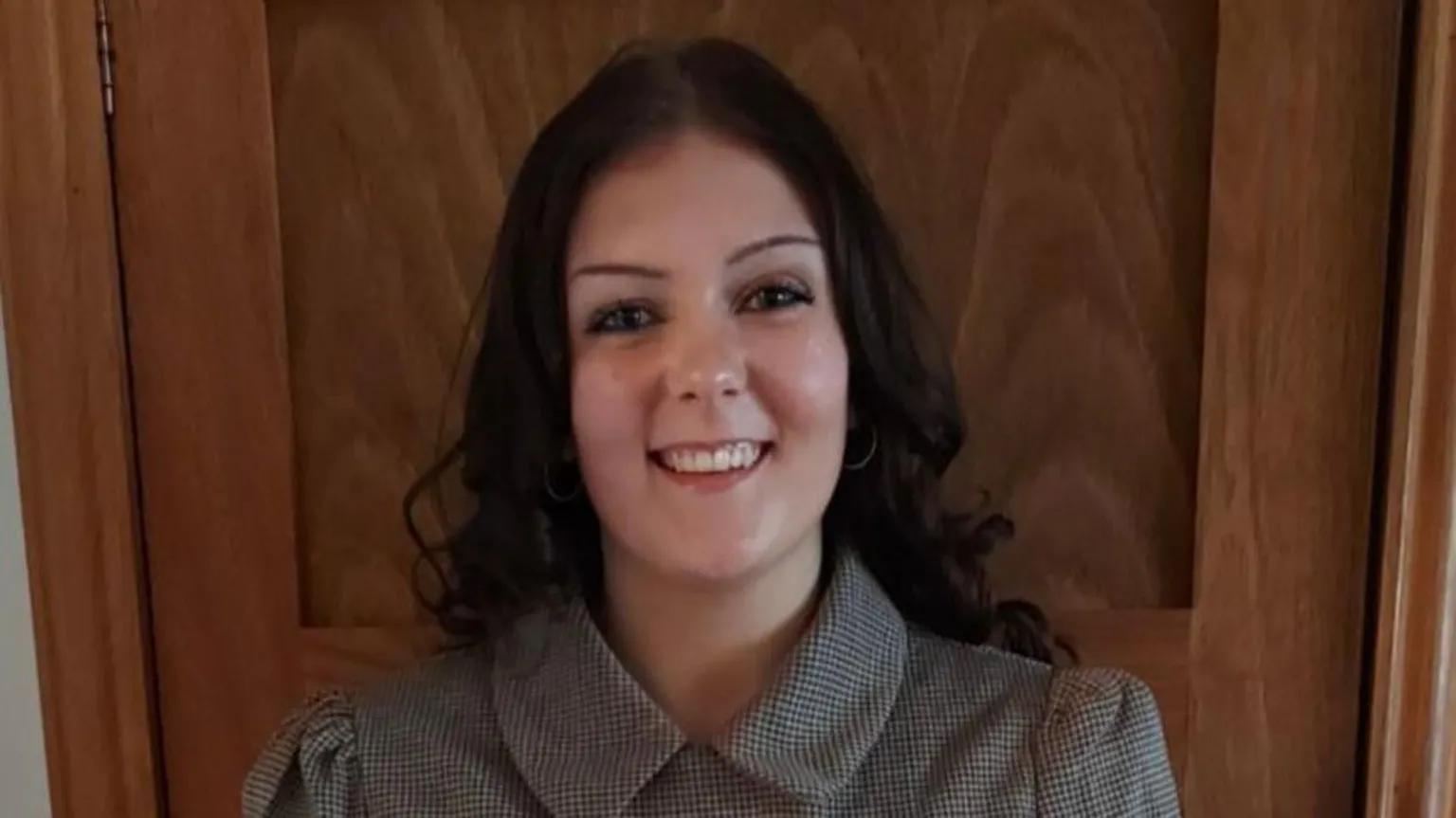 Darcy Ford Darcy Ford has long, dark brown hair and is smiling at the camera. She is wearing a checked top with a collar and is standing in front of a wooden door