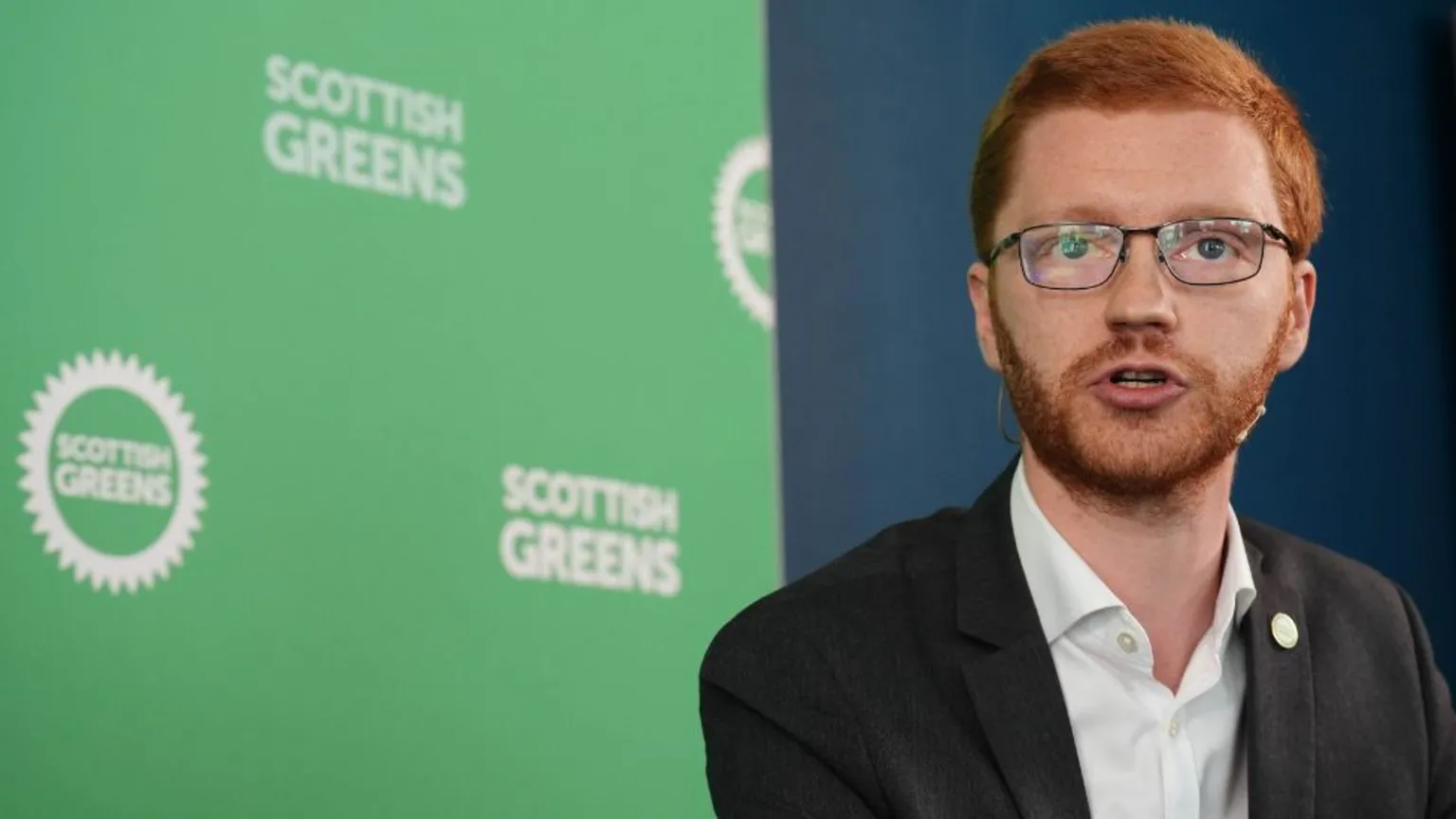 Ross Greer, who has ginger hair and glasses, pictured in front of Scottish Green branding. He is wearing a dark suit jacket and white shirt.