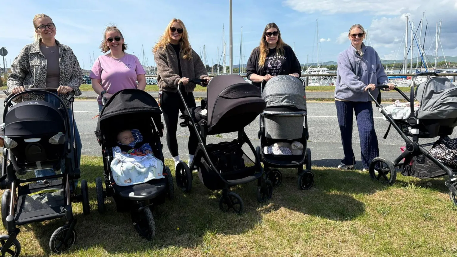 A group of mums with their strollers outside in the sunshine. The marina can be seen behind them.