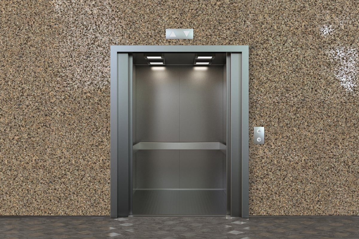 Empty elevator cabin with open doors: it is metallic silver inside and set in a pale brown stone wall.