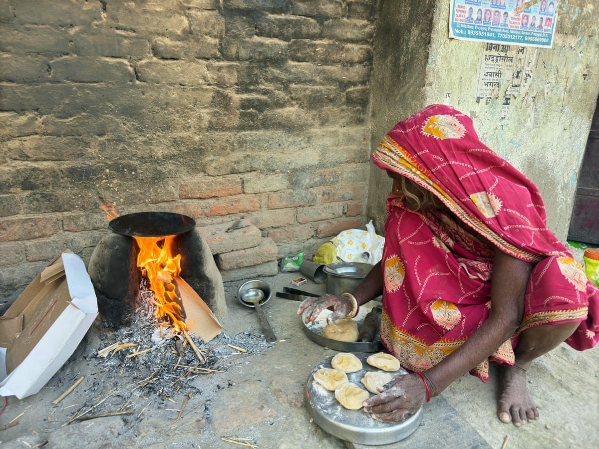 A woman draped in a bright pink and gold sari and scarf over her head crouches beside a small fire with a tray of patties.