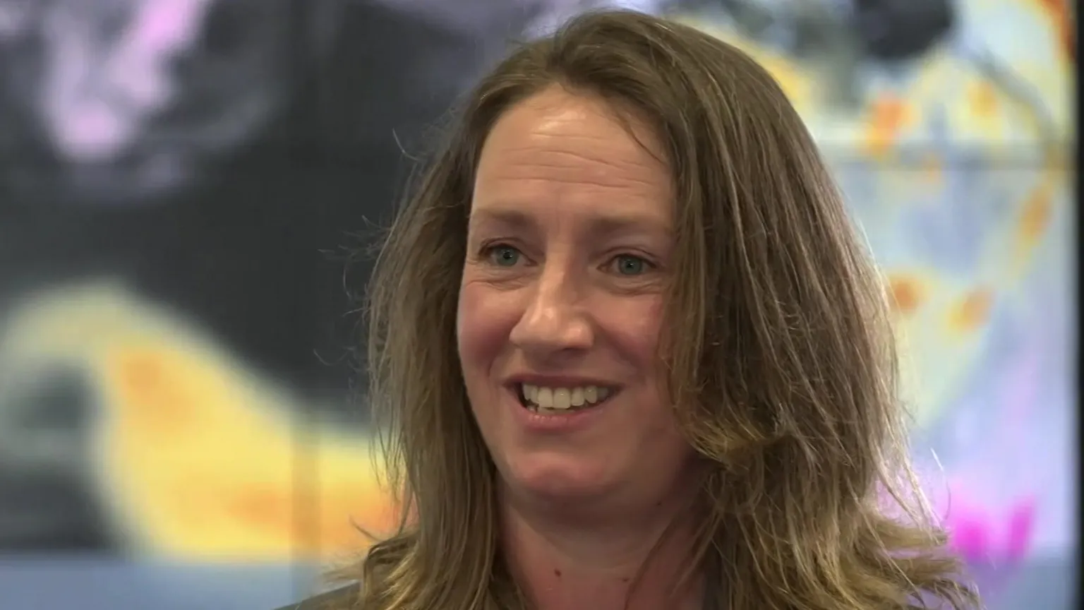 A picture of Melanie Ades, who has brown shoulder length hair and is stood in front of a large screen behind her.
