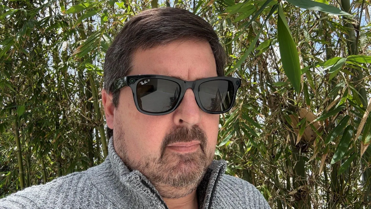 Mark Smith A man standing outside in a grey sweater and wearing Meta's Ray-Bans with a wall of green bamboo behind him.