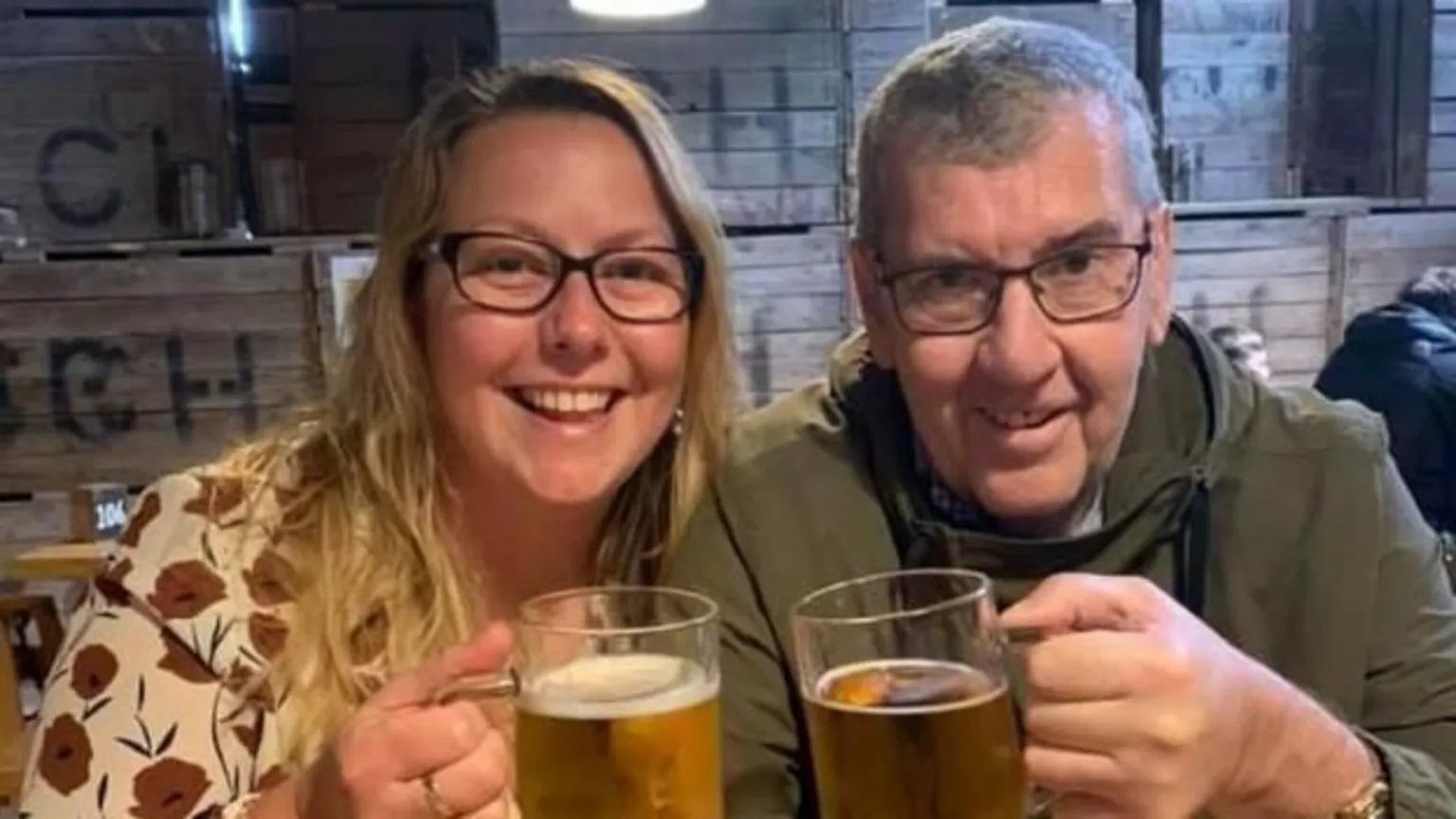 Blonde woman, Maureen, and grey-haired man Tony Dynes, in a pub. Both are smiling at the camera and holding up pints of beer. Maureen is wearing a patterned cream top with red flowers and Tony is wearing a green jacket. Both are wearing glasses. 