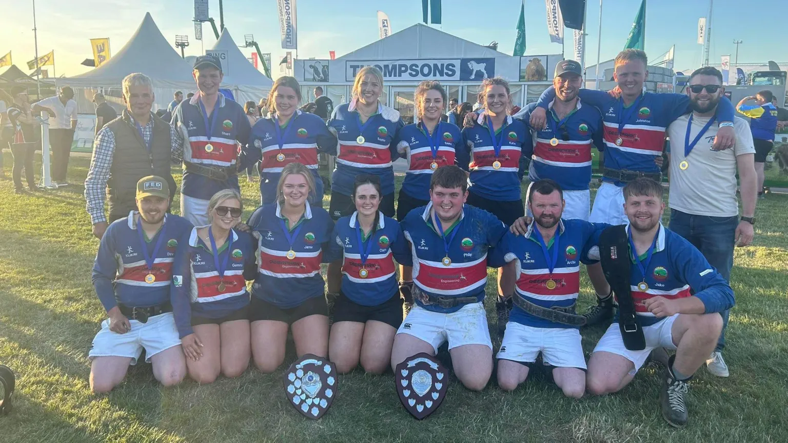 Lisnamurrican YFC Young men and women wearing blue and red sports jerseys pose in front of two trophies. 
