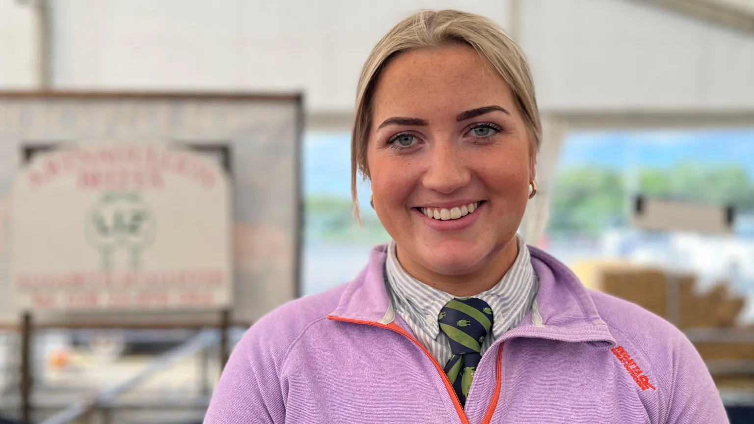 A woman with tied back blonde hair smiles into the camera. She is wearing a pink top over a white shirt and green and navy tie.