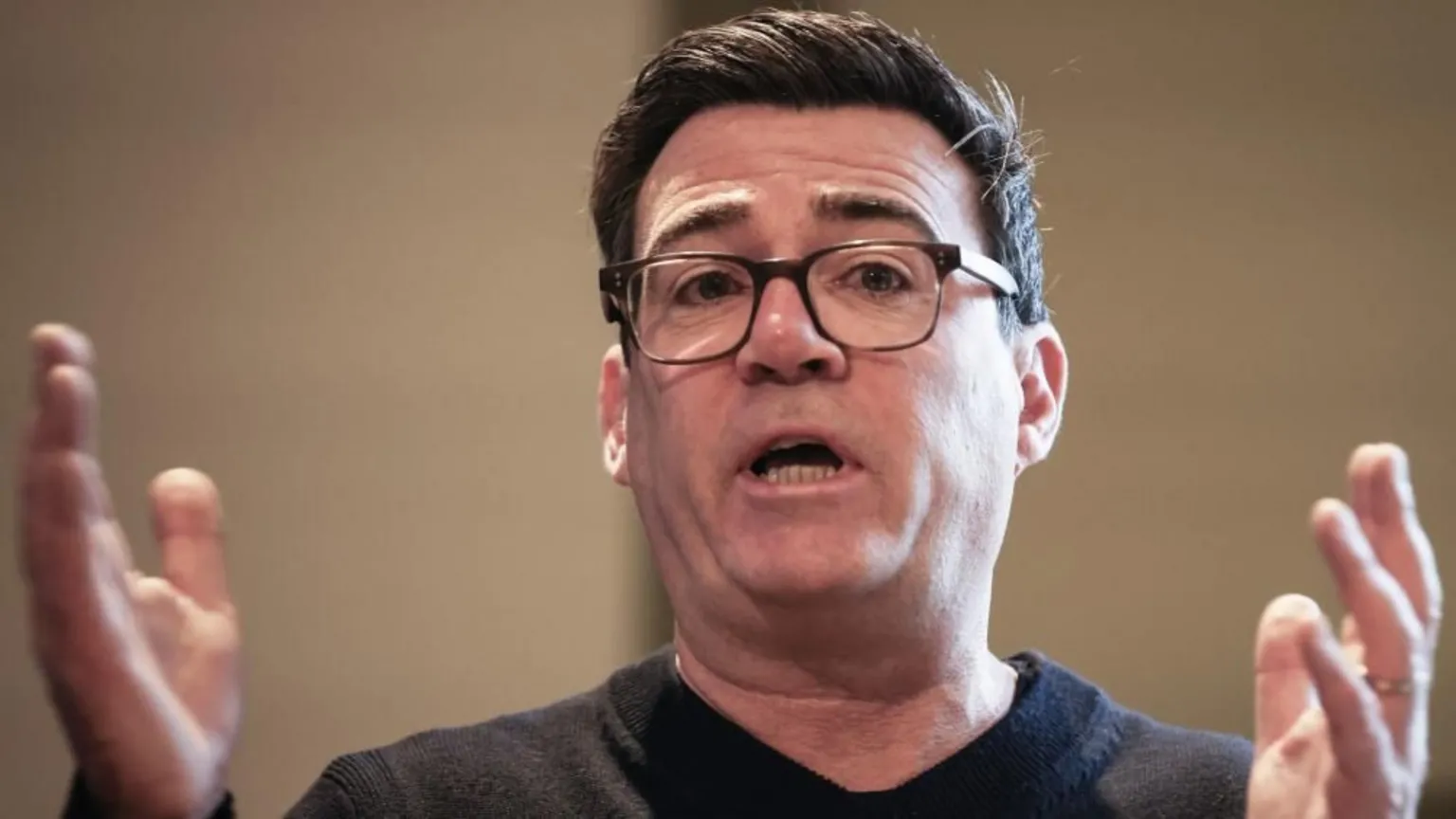  Andy Burnham is wearing a black jumper and black glasses. He is speaking with his hands raised and held apart.