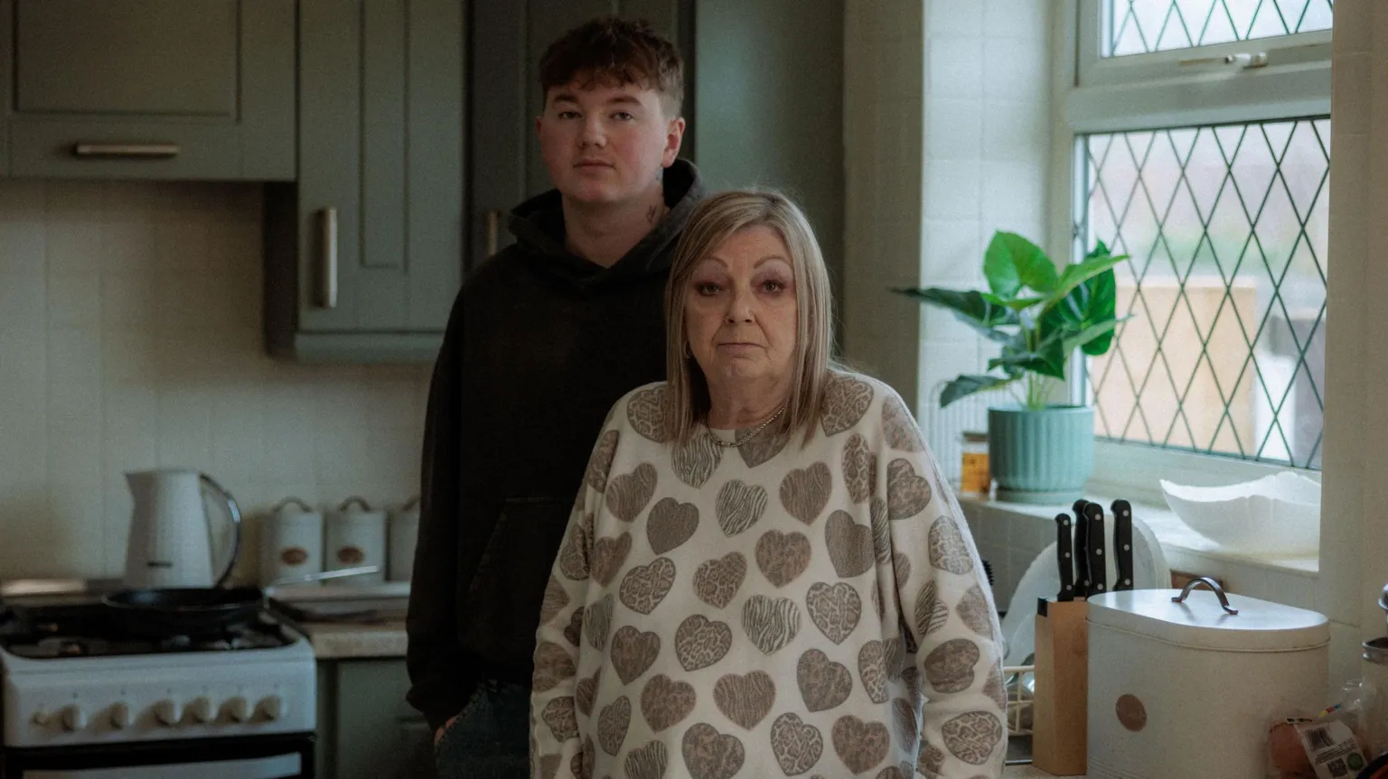 Alex Batty is standing next to his grandmother Susan Caruana in a kitchen with green cabinets and kitchen utensils on the counter. 