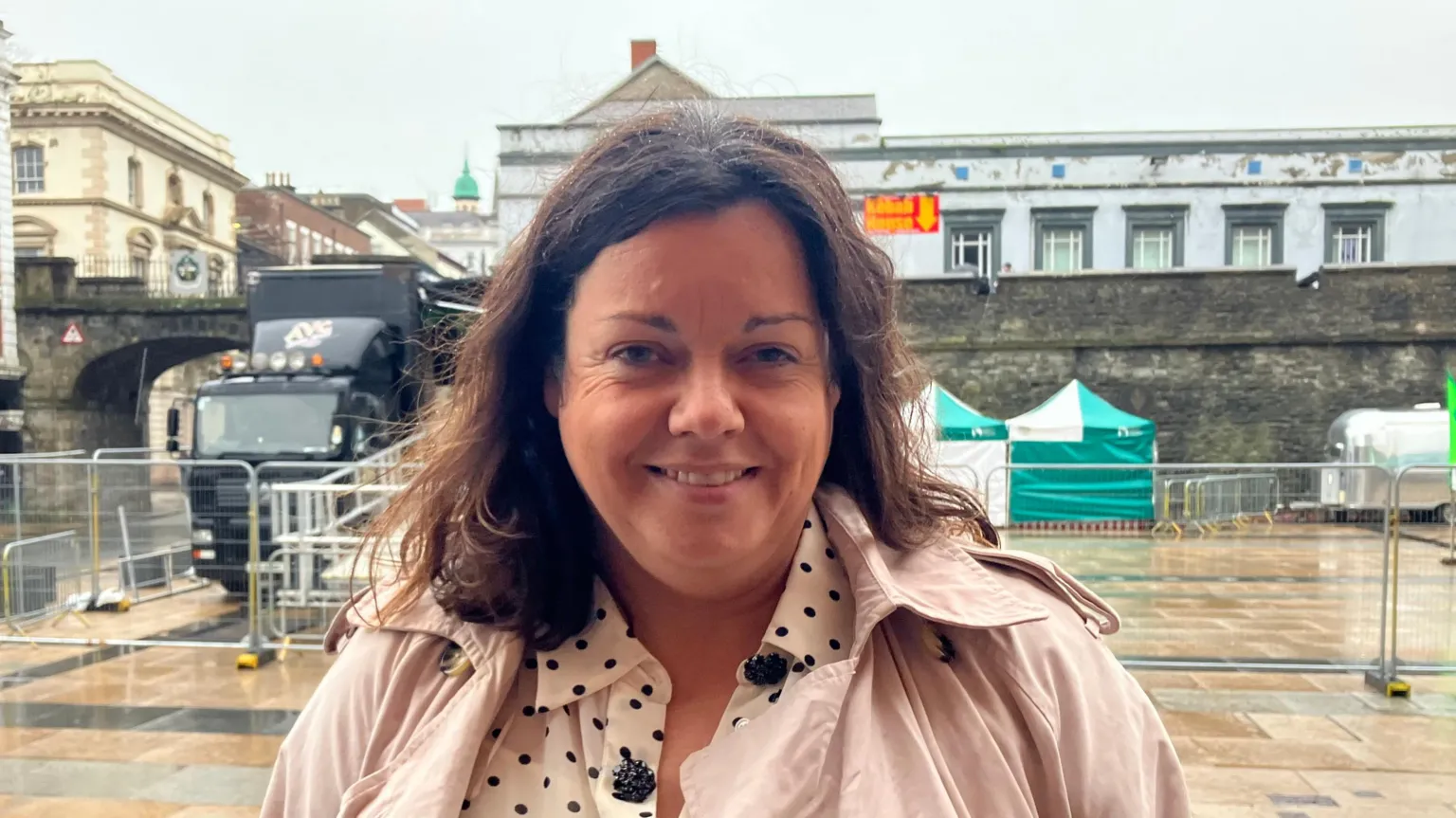 Jacqueline Whoriskey is wearing a pink jacket, and black and white shirt. She is standing in Guildhall Square with a number of stalls and a black lorry visible in the background.