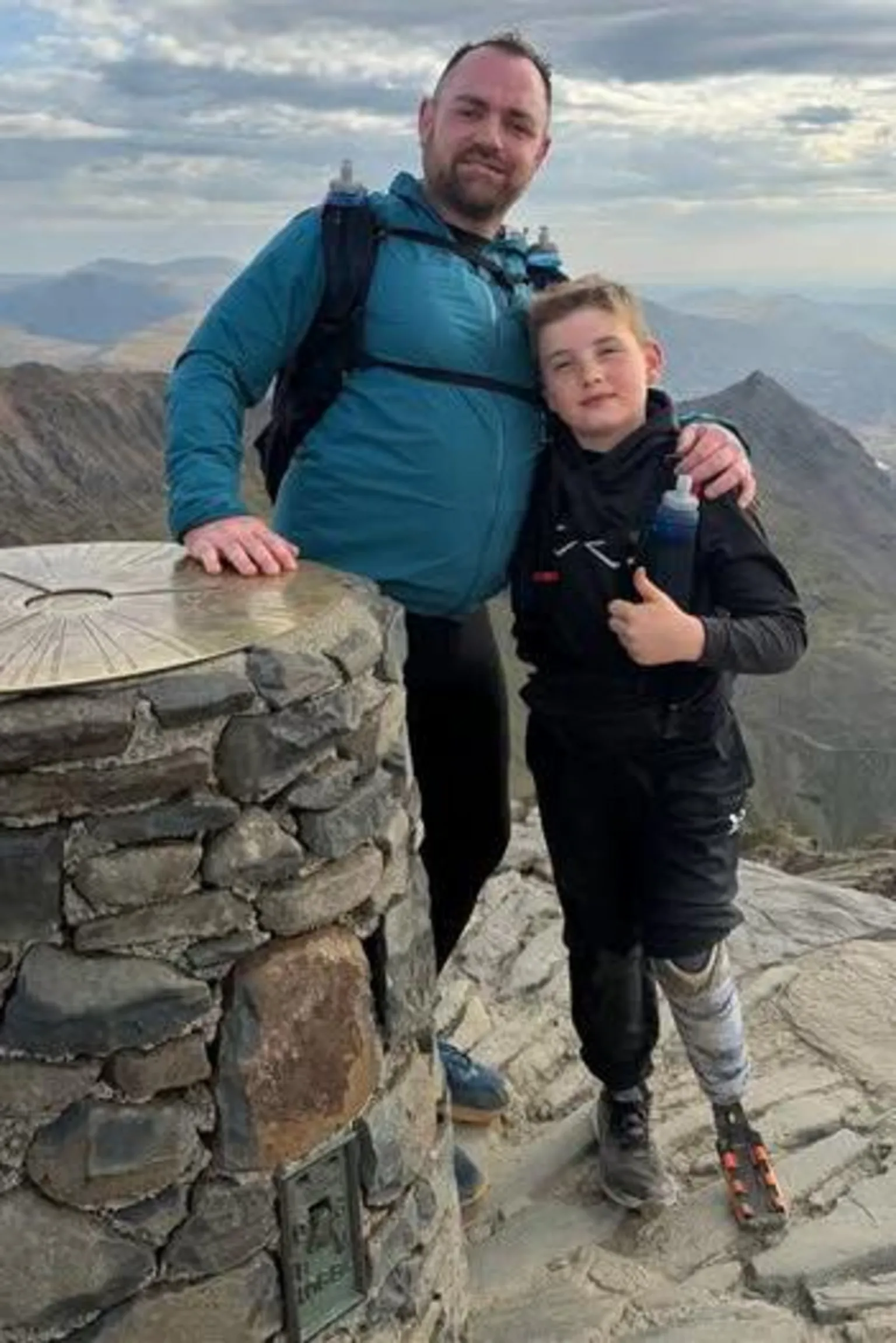 Daniel Thomas A man in his 40s with short dark hair and a short dark beard and wearing a blue waterproof coat stands at the summit of the mountain Yr Wyddfa with his arm around a boy of 9 who is wearing a black coat and black trousers, he also has a prosthetic leg on the left side. Both are wearing rucksacks and the boy is giving a thumbs up to the camera. The sky behind is a mix of blue and cloud and the mountains are visible stretching into the distance.