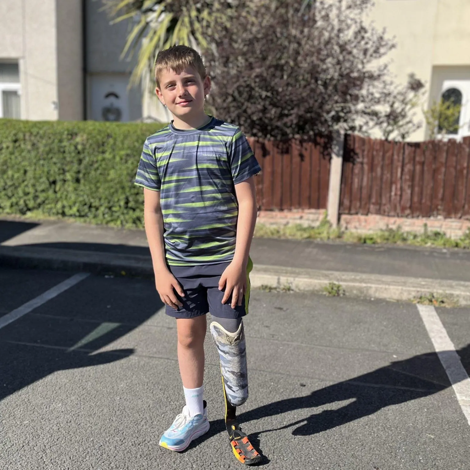 A boy stands on a residential street in the sunshine with two cream coloured houses in the background and a large bush behind a brown fence. He is smiling at the camera and wearing a grey and yellow patterned sports top and grey shorts. He has a prosthetic blade on his left leg and on his right he is wearing a blue and white trainer.