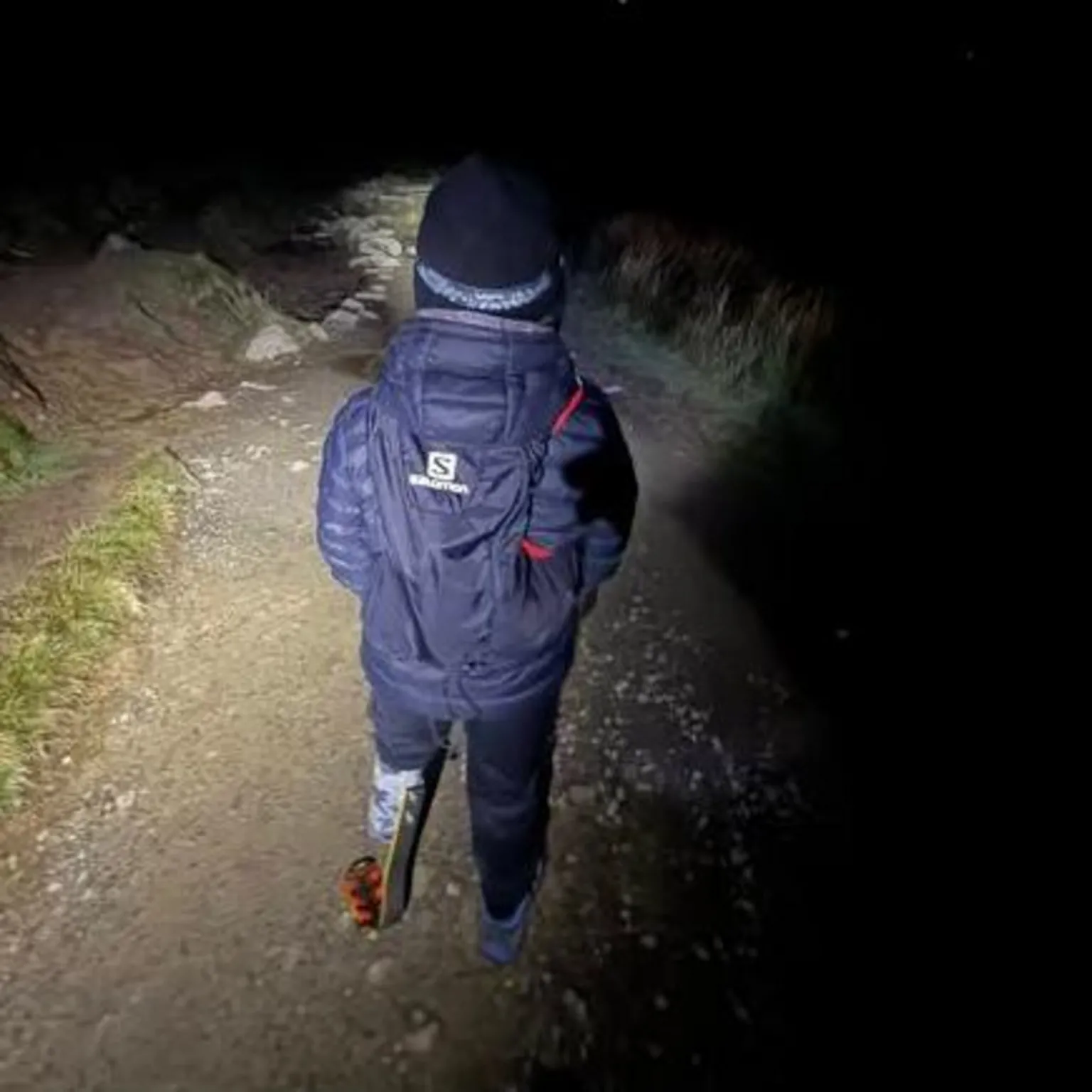 Daniel Thomas A picture of a young boy with a prosthetic limb on the left side on a mountain path in the dark. The image is taken from behind. He is wearing a navy padded coat and a black hat and the way in front of him is lit by a head torch that he is wearing. He is also carrying a navy rucksack.