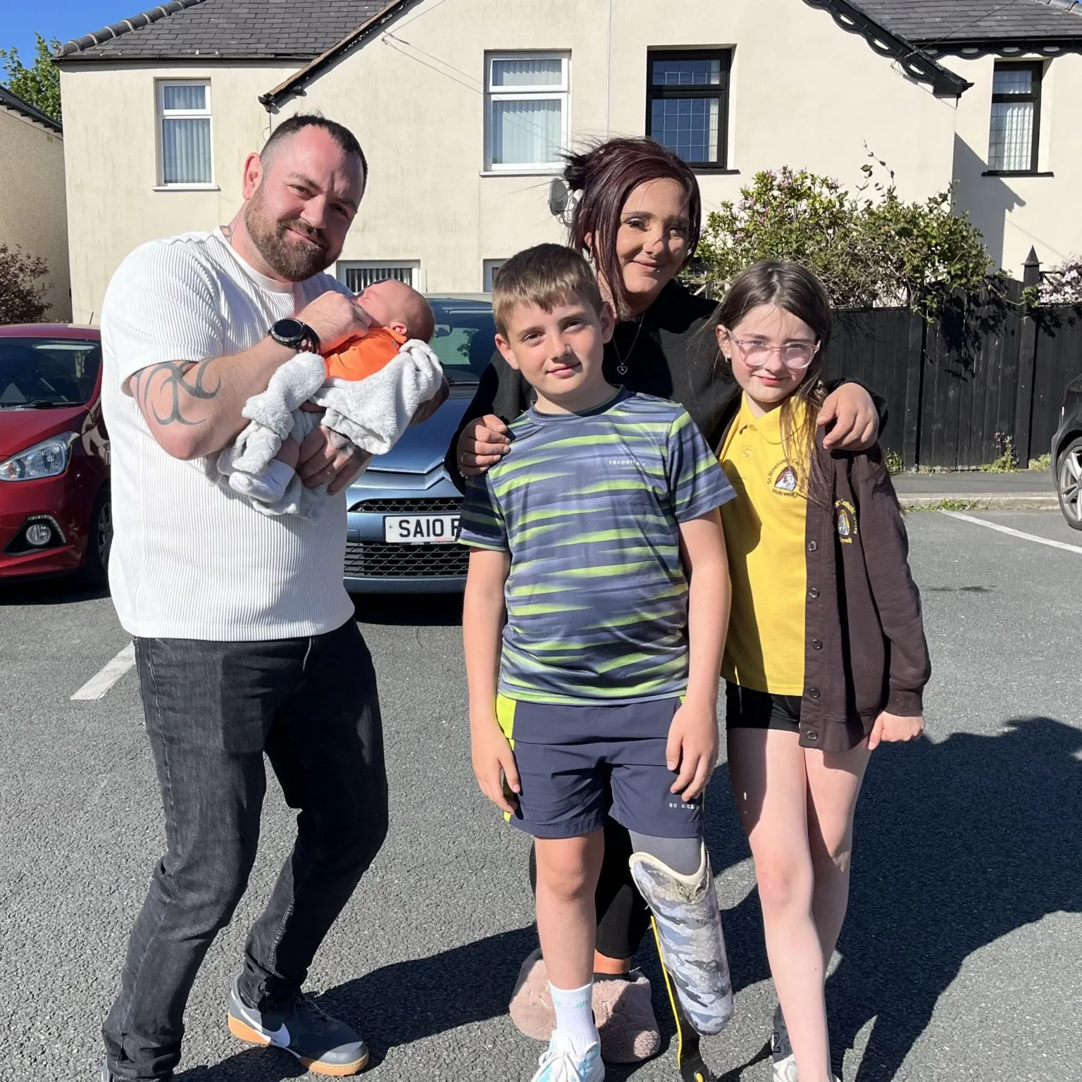 A picture of a mum and dad on a sunny street with cream semi-detached houses in the background. The man, with dark hair and a short dark brown beard is wearing jeans and a white t-shirt, and is holding a newborn baby and the woman, who has dark hair tied up, is standing behind two children, a boy and a girl, with her hands on their shoulders. The boy has a prosthetic leg and is wearing shorts and a t-shirt and the girl, who has long brown hair and glasses, is wearing a school polo shirt, cardigan and shorts.