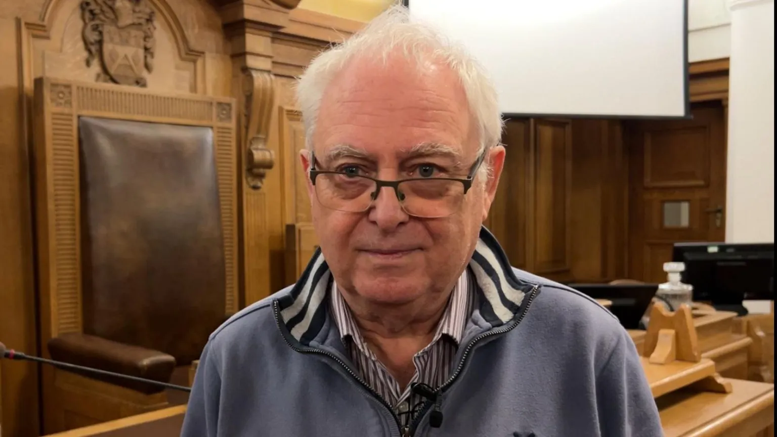 Steve Hey has short white hair and glasses. He wears an unzipped blue fleece top and a striped shirt. He stands in the council chamber.
