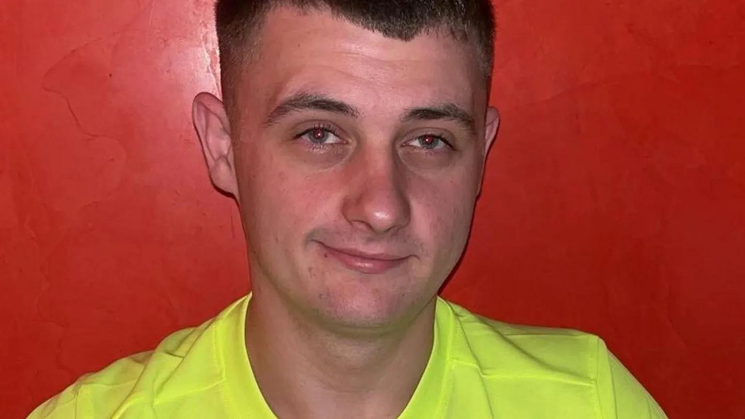 Spindrift Fraser Stewart is half smiling at the camera. He has short, brown hair, shaved at the sides and is wearing a luminous yellow T-shirt. He is standing against a red wall.