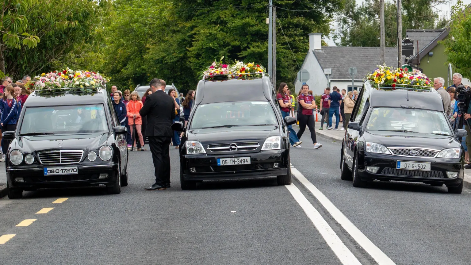  Three funeral hearses driving on a road with flowers on top of them. Crowds of people are behind it.