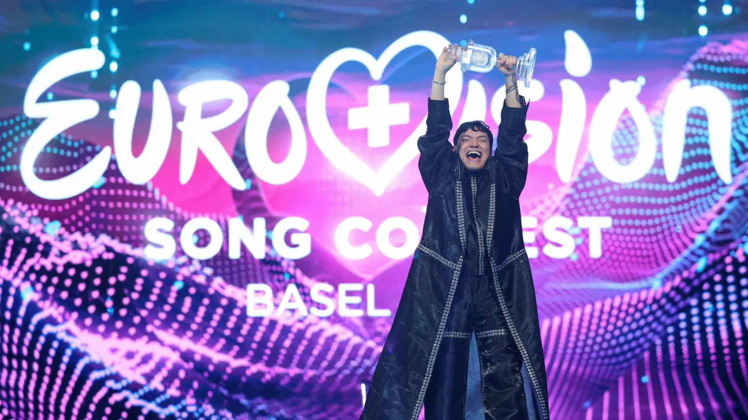  JJ holding aloft the trophy in celebration at Eurovision 2025 in front of a giant colourful Eurovision Song Contest branded backdrop