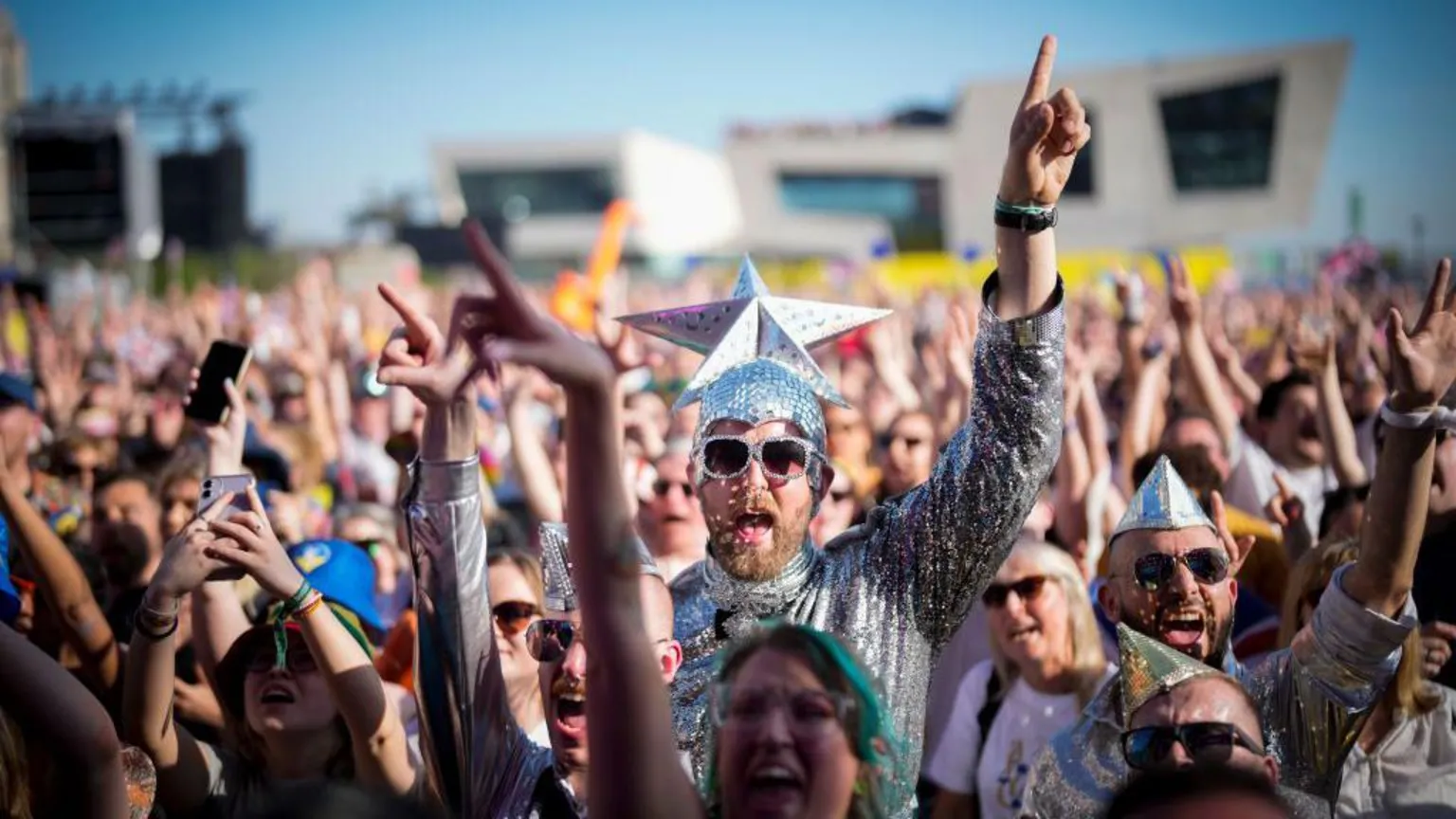  Eurovision fans watch the 2023 Eurovision Song Contest final on a giant screen in host city Liverpool. Many fans are dressed up, including the man in the centre of the photo who wears a silver jumpsuit and sunglasses and wears a silver cap with a giant silver star on the top.