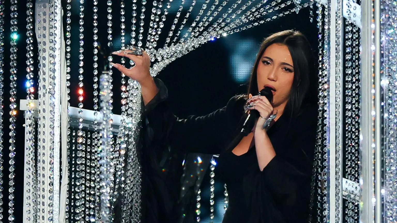  Yuval Raphael sings on stage, wearing a black dress and jacket. She holds open a glass beaded curtain. 