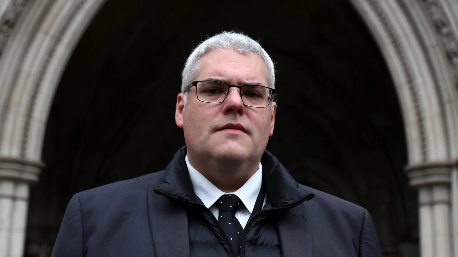  Gavin Robinson is looking down the barrel of the camera. Hew has grey hair and black glasses. He is wearing a white shirt, black tie and black coat.