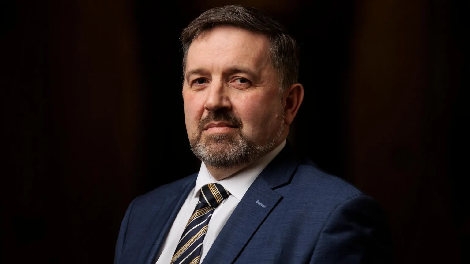  Robin Swann is looking directly at the camera. He has black hair and a grey beard. He is wearing a white shirt, stripe tie and navy blazer. 