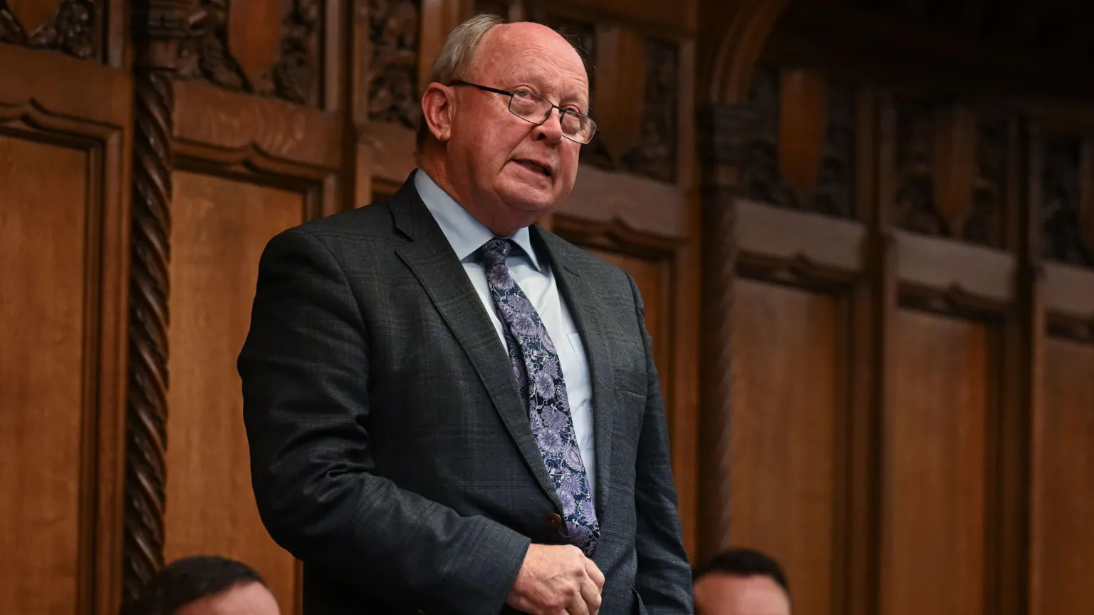  Jim Allister is speaking in the House of Commons. He is standing up with a brown wood panelled wall behind him. He is wearing glasses, a grey suit, white shirt and floral tie. 