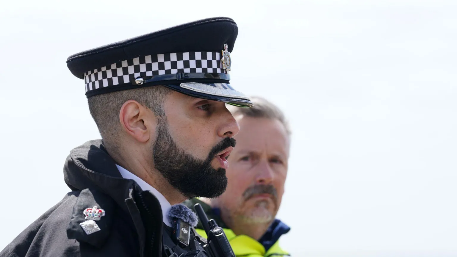 Side profile of a male police officer with black beard. To his left is a man in a high-vis jacket.