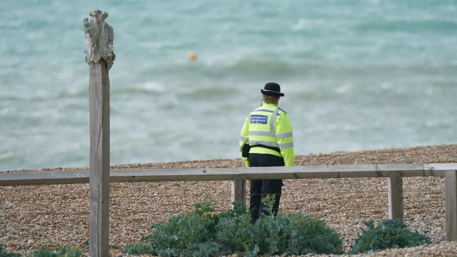 A lone police officer stood on a beach facing away from the camera towards the sea