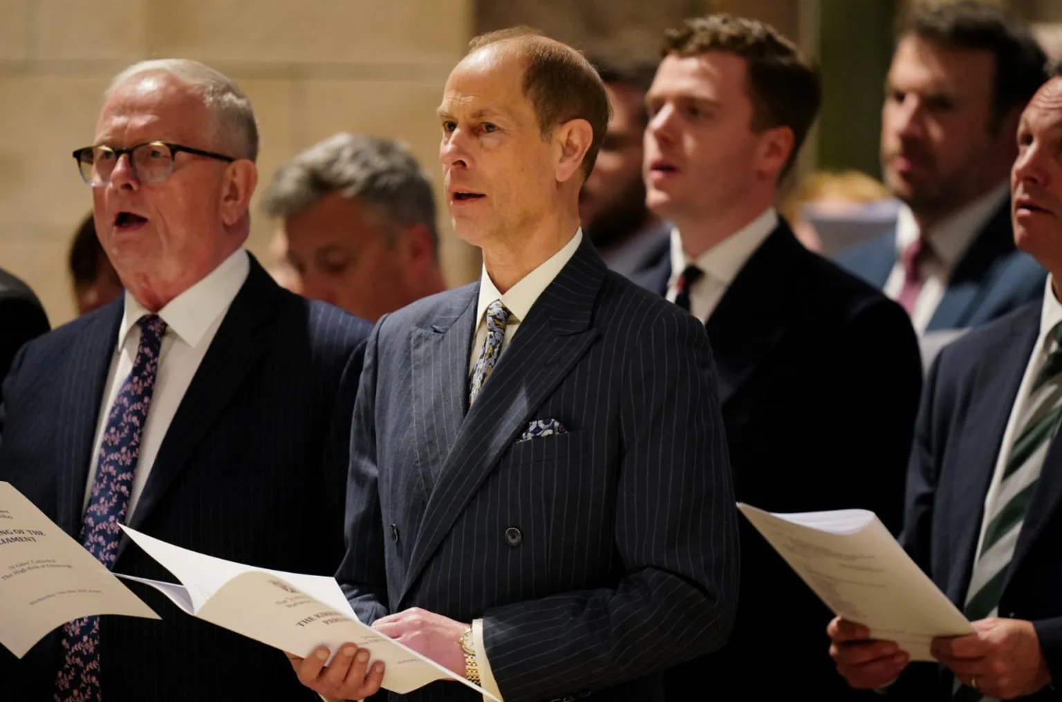 Men in suits stand singing from printed hymn sheets during. The central man wears a dark pinstripe double-breasted suit with a patterned tie and pocket square. Several other men are visible behind him, also holding service booklets and singing. The setting has warm lighting and stone architecture, giving a ceremonial atmosphere.