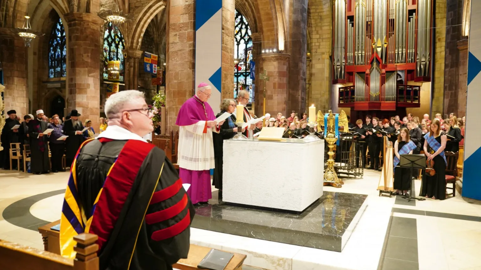 Clergy members lead a ceremonial service at the altar inside St Giles' Cathedral in Edinburgh during the Royal Kirking of Parliament. Choir members and musicians stand near a large pipe organ in the background, while representatives from different faiths and civic groups hold programmes along the side aisle. A man in ceremonial academic robes sits in the foreground beneath the cathedral’s stone arches and stained glass windows.