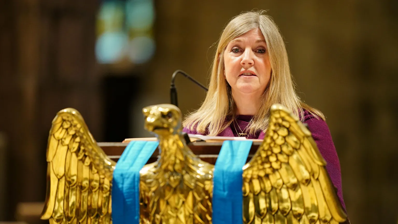 woman with blonde hair stands behind a gold eagle statue as she reads in churh