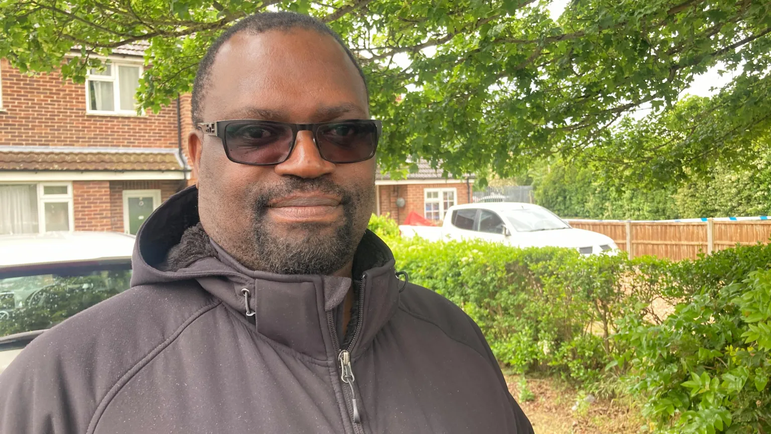 Mousumi Bakshi/BBC A man wearing a black coat and sunglasses is stood on a driveway with a house and greenery behind him