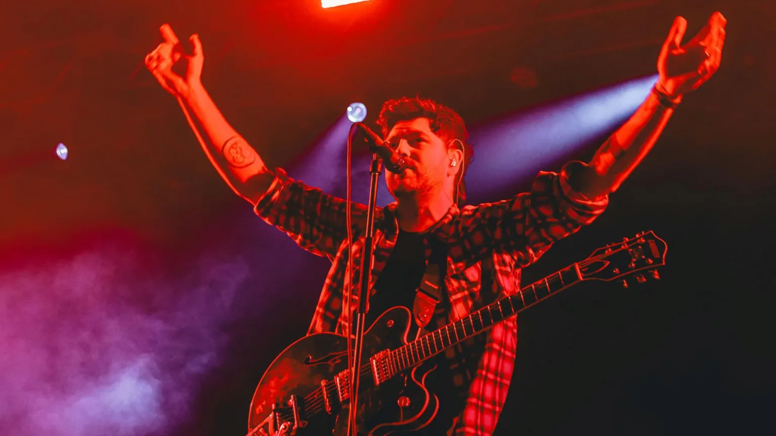 Twin Atlantic frontman Sam McTrusty, pictured at the Y Not Festival in 2023, is standing on a stage with a guitar slung over his shoulders and his arms raised. The musician is bathed in a red glow.