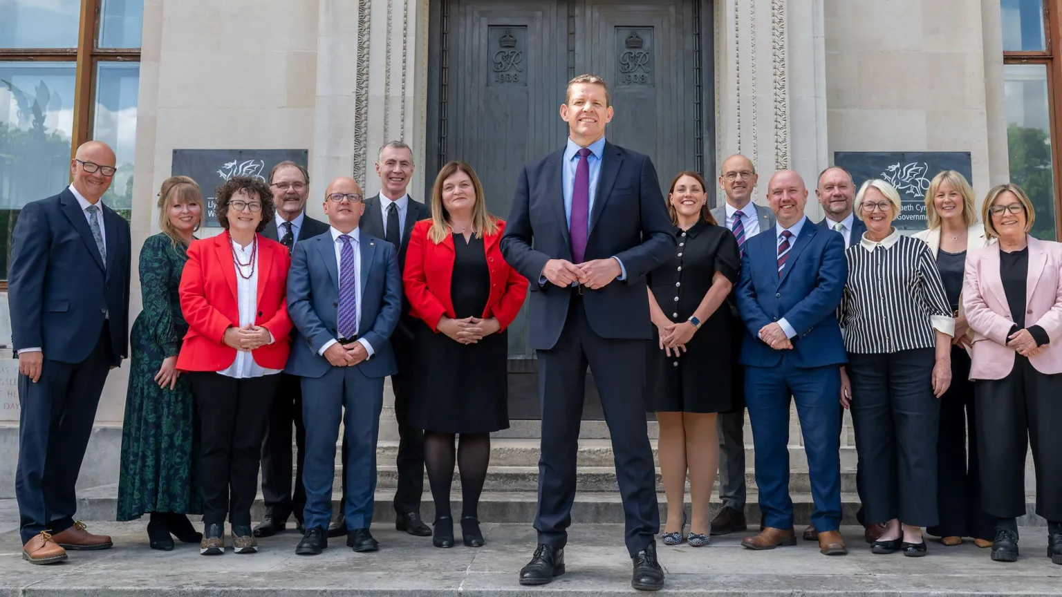 Welsh government The first minister and cabinet ministers stand on steps in front of a grey building with a brown door.
