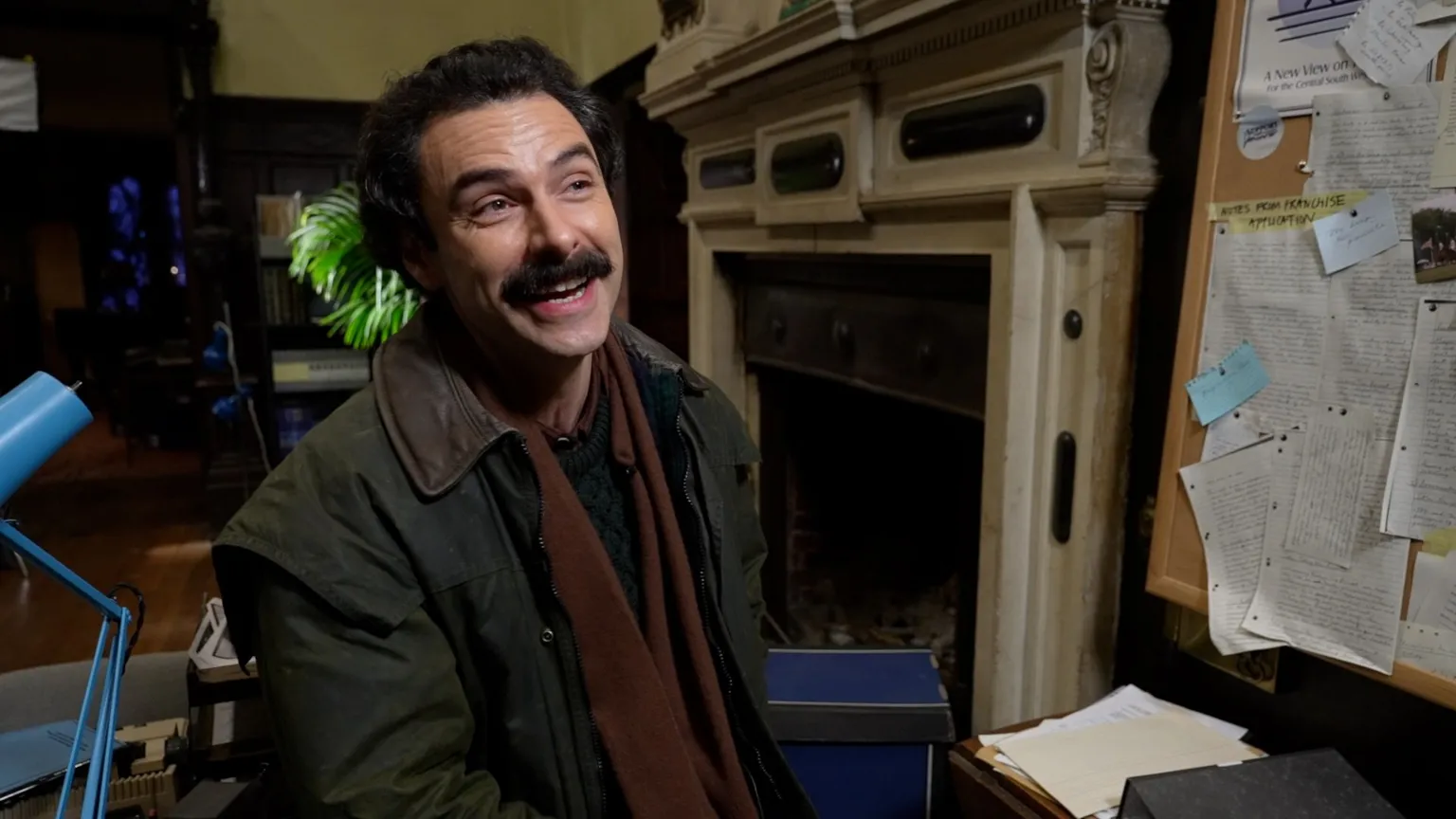 Julius Peacock/BBC Aiden Turner laughs as he speaks to someone off camera. He's wearing a green jacket with brown scarf, and has a thick moustache. 
