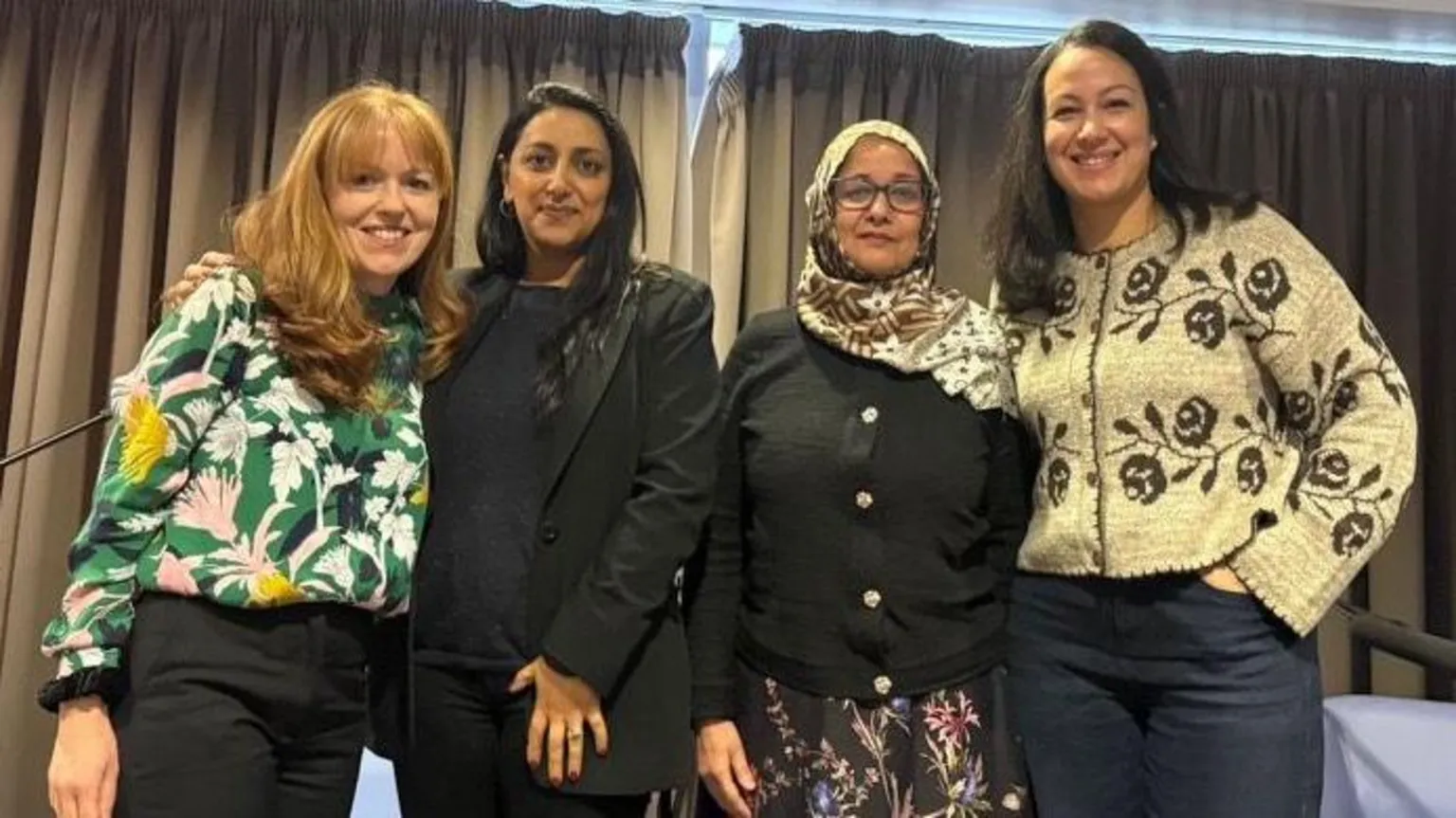 Four women stand closely together, smiling at the camera, after a panel discussion. Behind them are four chairs and banners for BBC Cymru Wales and Muslim Doctors Cymru.