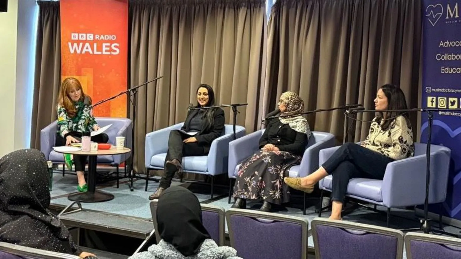Four women are seated on a stage, with microphones in front of them, as they talk to an audience. In the foreground are rows of seats with two women in the audience. On the stage behind the women are banners for BBC Radio Wales and Muslim Doctors Cymru.