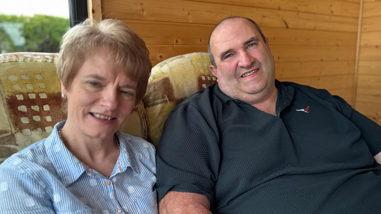 Marshall, a big man in a dark polo shirt, sits beside a woman in a pin-stripped shirt on a sofa. Behind them is a wood panelled interior wall. 