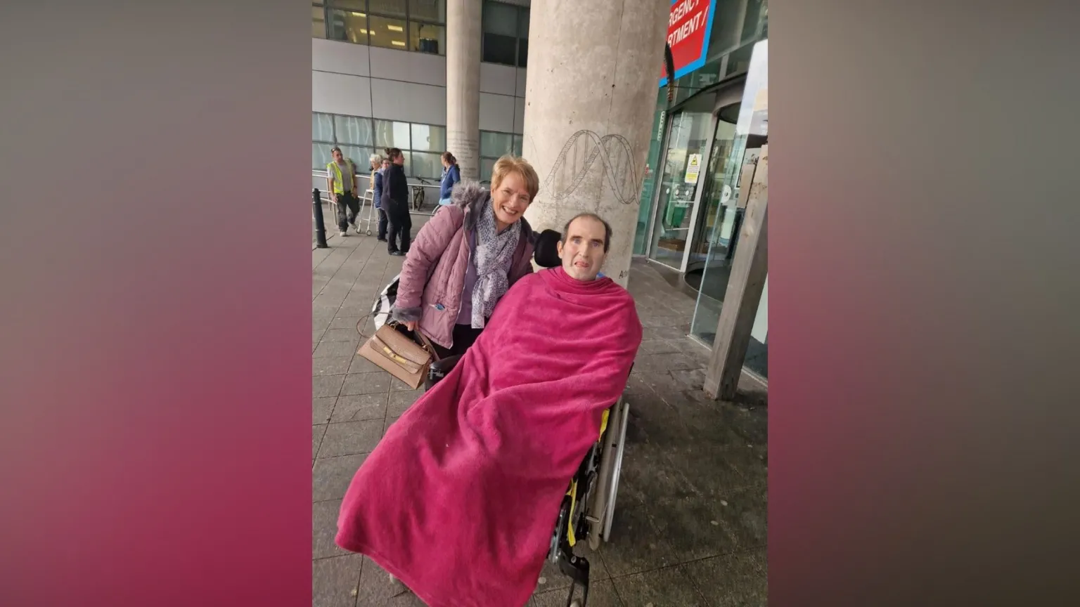 Marshall Wylie Marshall is in a wheelchair and is wrapped a pink blanket. He is outside a hospital and his wife is standing behind him. 