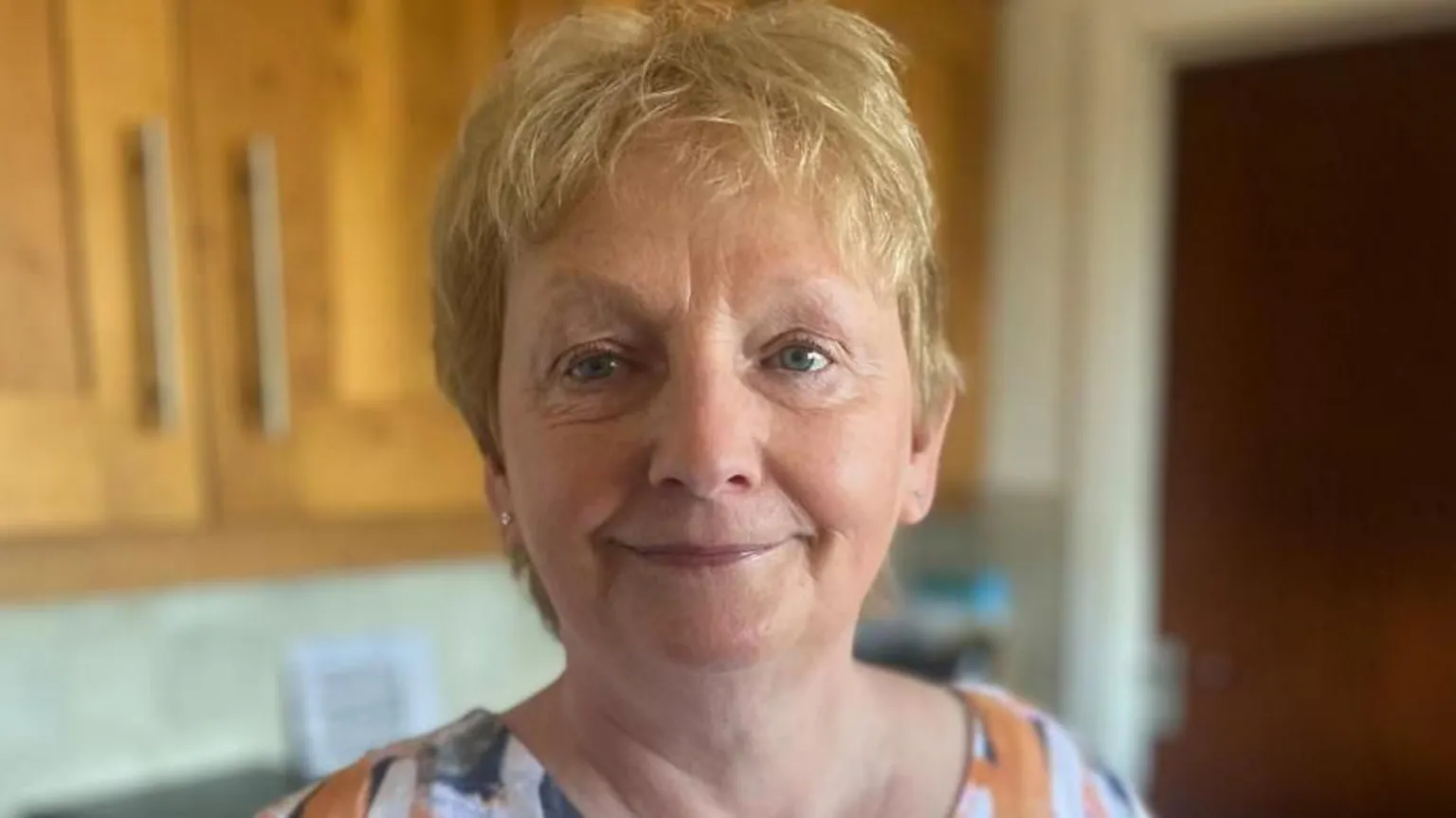 Freda is smiling at the camera. She has short ginger hair and is wearing a orange patterned top.